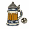Épingle en émail souple représentant une chope de bière bavaroise avec couvercle, garniture en métal argenté, bandes à carreaux bleues, émail ambré pour la bière et pochette papillon sur fond blanc.