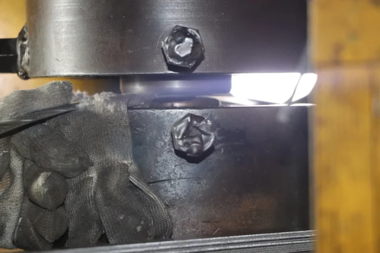 A close-up of a worker wearing protective gloves operating a metal stamping machine at the exact moment of pressing, showing the industrial production process in a factory setting.
