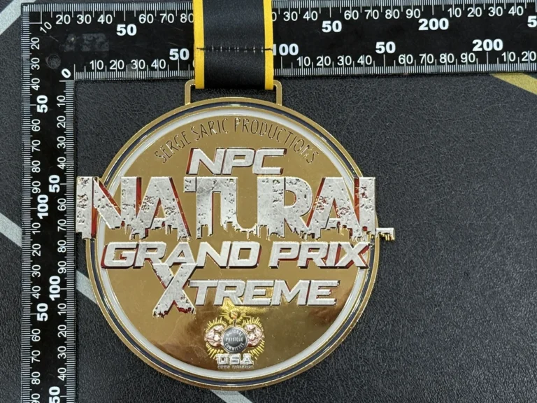 NPC Natural Grand Prix Xtreme용 원형 금메달의 클로즈업으로, 각인된 행사 문구, 은색 포인트, 그리고 검은 리본이 특징입니다.
