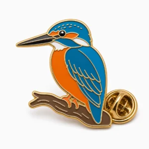 Spilla a forma di martin pescatore appollaiato con contorni in rilievo in metallo dorato, dorso blu, ventre arancione, gola bianca, ramo marrone e frizione a farfalla su sfondo bianco.
