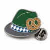 Spilla per cappello bavarese a tesa pretzel con contorni in metallo argentato lucido, cappello verde, dettaglio pretzel abbronzato, fascia bianca blu e frizione a farfalla su sfondo bianco.