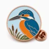 Épingle représentant un martin-pêcheur, avec des contours en métal doré rose, une base ronde bleu pâle, une tête bleue, une joue orange, des accents de roseau et une pochette de papillon sur fond blanc.
