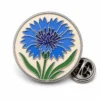 Spilla floreale rotonda in fiordaliso con contorni in metallo argentato lucido, base circolare color crema, fiore blu vivo, foglie verdi e una farfalla in frizione su sfondo bianco.