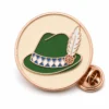 Spilla rotonda per cappello bavarese dell'Oktoberfest con contorni in metallo oro rosa, base rotonda color crema, cappello verde, fascia bianca blu e farfalla su sfondo bianco.