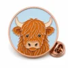 Round portrait Highland cow pin with rose gold metal outlines, pale blue base, orange-brown shaggy face, cream horns, and a butterfly clutch on white.