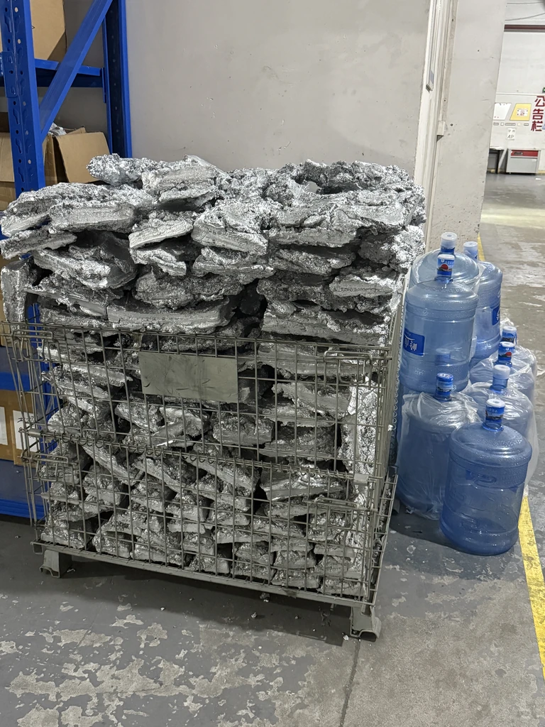 A metal cage filled with leftover zinc alloy pieces, representing the scrap material collected after the die casting process, placed in a warehouse environment next to water bottles.