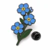 Épingle "forget me not" en forme de grappe avec contours en nickel noir, fleurs connectées en émail bleu, centres jaunes, tige verte, et une pochette papillon sur fond blanc.