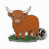 Standing Highland cow pin with polished silver metal outlines, rust brown shaggy coat, cream horns, green grass base, and a butterfly clutch on white.
