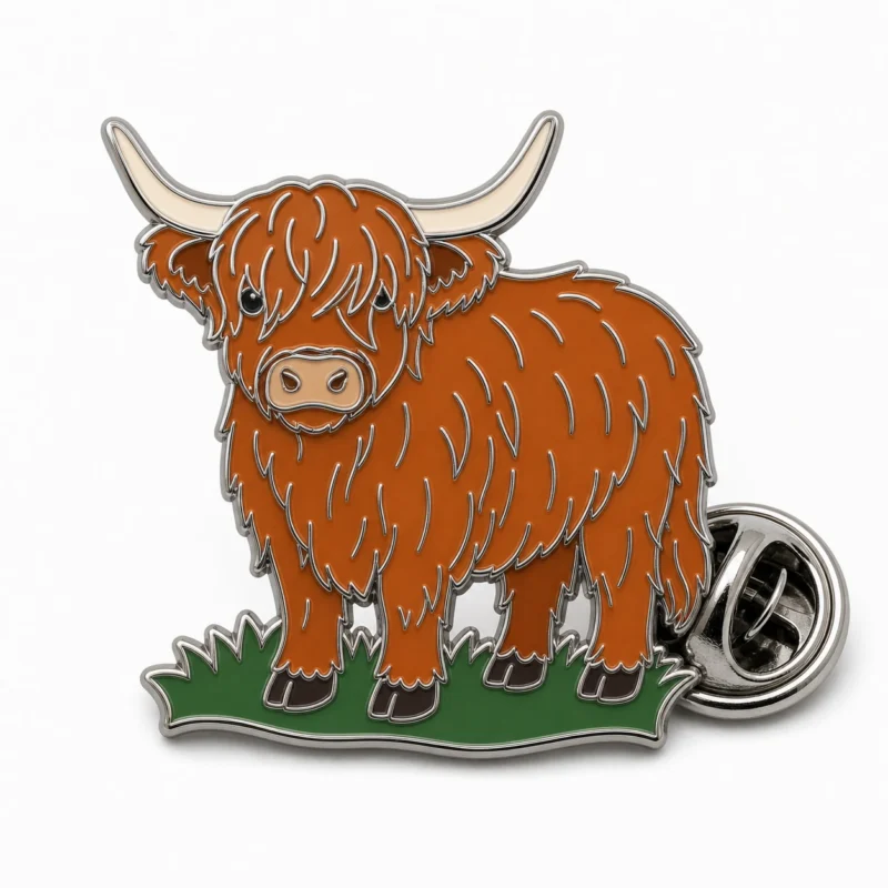 Standing Highland cow pin with polished silver metal outlines, rust brown shaggy coat, cream horns, green grass base, and a butterfly clutch on white.