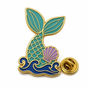 Spilla a forma di coda di sirena verde acqua con contorni in rilievo in metallo dorato, forma di coda verde acqua collegata, accento di conchiglia viola, base a onda blu e frizione a farfalla su sfondo bianco.