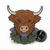 Thistle Highland cow pin with black nickel outlines, rust brown shaggy face, cream horns, purple thistles, green leaves, and a butterfly clutch on white.