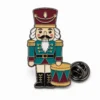 Alfiler cascanueces de tambor de juguete con contornos de níquel negro, sombrero burdeos, chaqueta verde azulado, cara crema, tambor dorado y un embrague de mariposa sobre blanco.