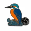 Épingle de martin-pêcheur à branches ondulées avec contours en nickel noir, aile bleu foncé, tête turquoise, ventre orange, base incurvée, et une pochette de papillon sur fond blanc.