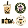 Значки с логотипом «Boba Tea»: эмалевые значки с изображением стаканчика для напитка, круглый значок «Boba Time», значок с буквой «Boba» и значок магазина с волнистым краем, выполненные в золотых и чёрных тонах на белом фоне.