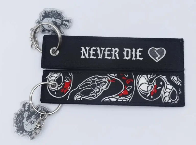 Two black embroidered fabric keychains with silver metal rings, one featuring “NEVER DIE” text and a broken heart, and the other detailed skeletal artwork with red accents.