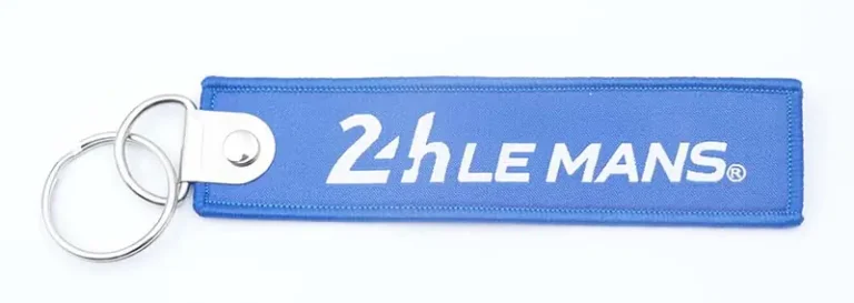 A blue woven fabric keychain with white logo text, stitched edges, silver metal key ring, and white leather-style connector tab.