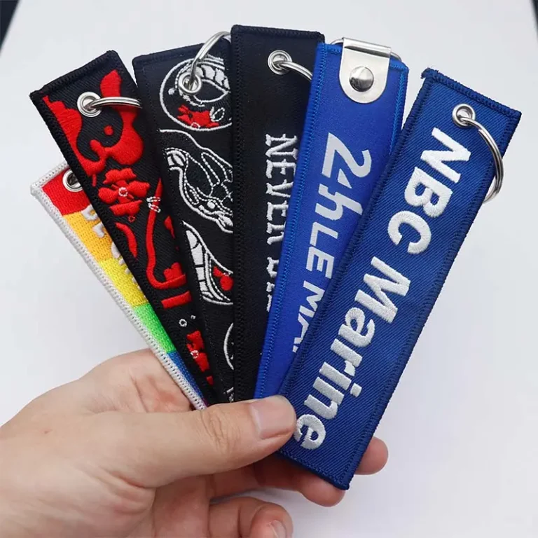 A hand holding several custom embroidered fabric keychains in black, blue, red, and multicolor designs, featuring stitched logos, text, metal eyelets, and silver key rings.