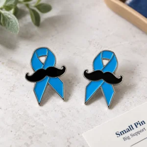 Two custom blue awareness ribbon enamel pins featuring black mustaches, polished silver-tone outlines, and a clean men’s health support design.