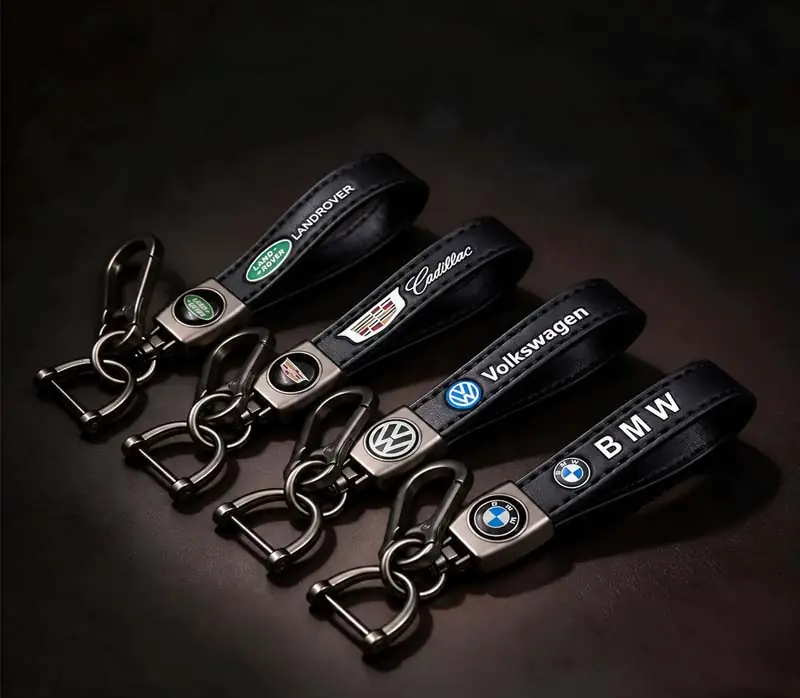 Cuatro llaveros de coche personalizados de cuero negro con los logotipos de Land Rover, Cadillac, Volkswagen y BMW, con correas de cuero cosidas, insignias metálicas con el logotipo, anillas para llaves y mosquetones en color gris plomo.